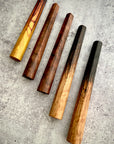 A collection of muddlers made from wood, displayed on a textured surface. They appear to be of varying sizes and have a rustic, wooden appearance with some having darker finishes.