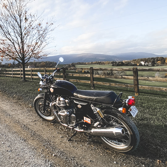 Royal Enfield INT650 motorcycle parked beside and fenced farm with mountains in background