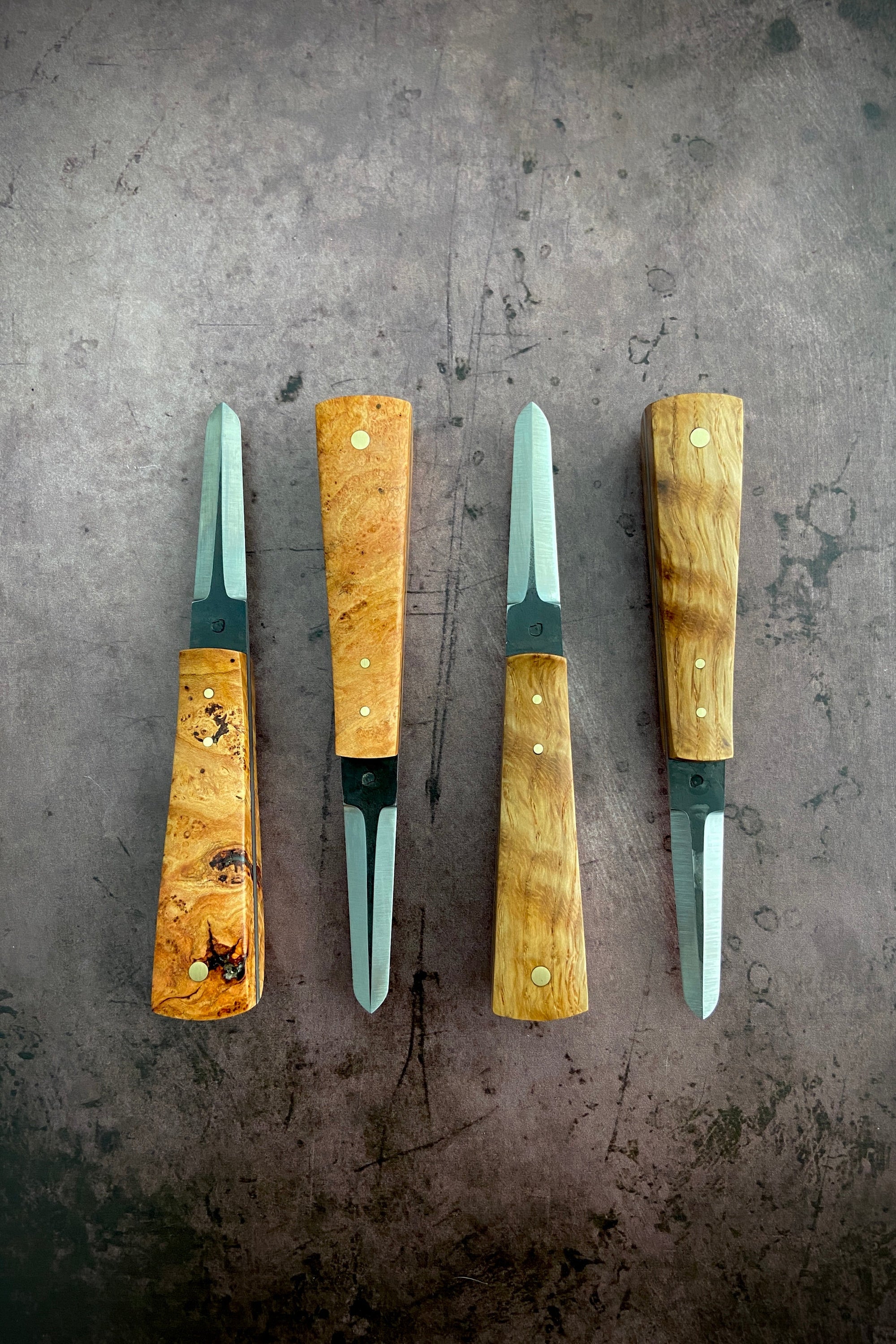 A set of four oyster knives with wooden handles and steel blades, displayed side by side on a textured metal surface.