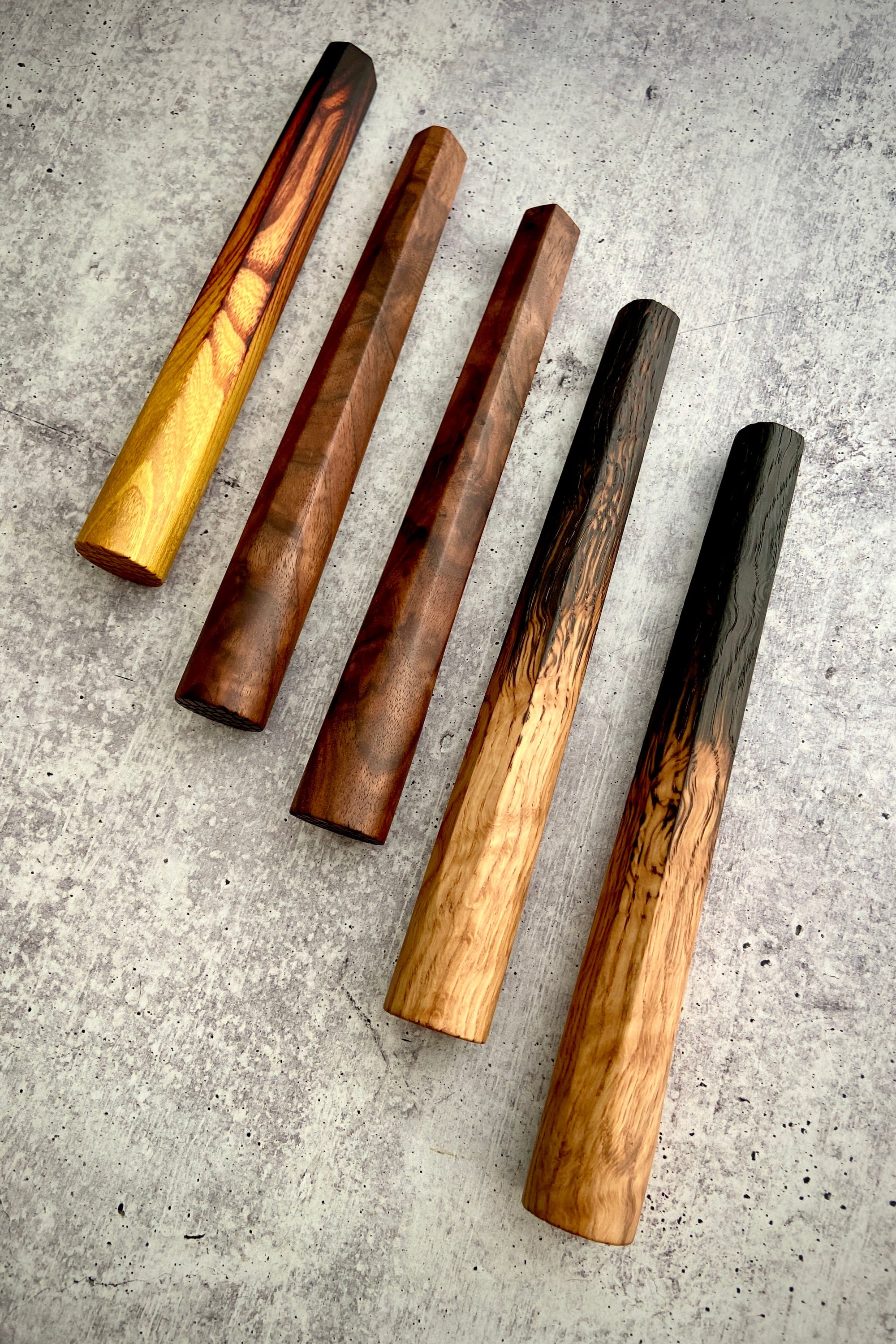 A collection of muddlers made from wood, displayed on a textured surface. They appear to be of varying sizes and have a rustic, wooden appearance with some having darker finishes.