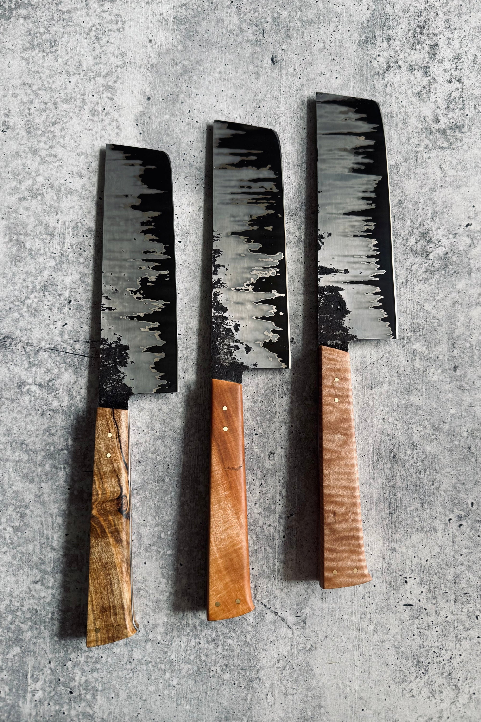 Three Japanese style vegetable knives with wooden handles and stainless steel blades, laid out on a textured grey surface.